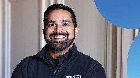 Oregon Executive MBA graduate Shan Rammah smiles while posing in front of an off-white wall. 